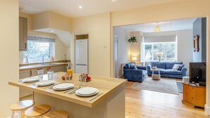 A modern kitchen with a central island, dining area with stools, a living room with a blue sofa, and a TV. - 3 Bed in Looe (oc-s29266) (Looe)