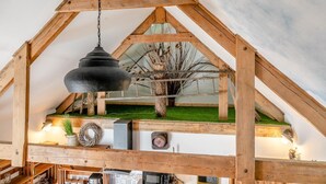 Un espacio tipo loft con vigas de madera a la vista, una lámpara colgante, un pequeño árbol y césped artificial verde.