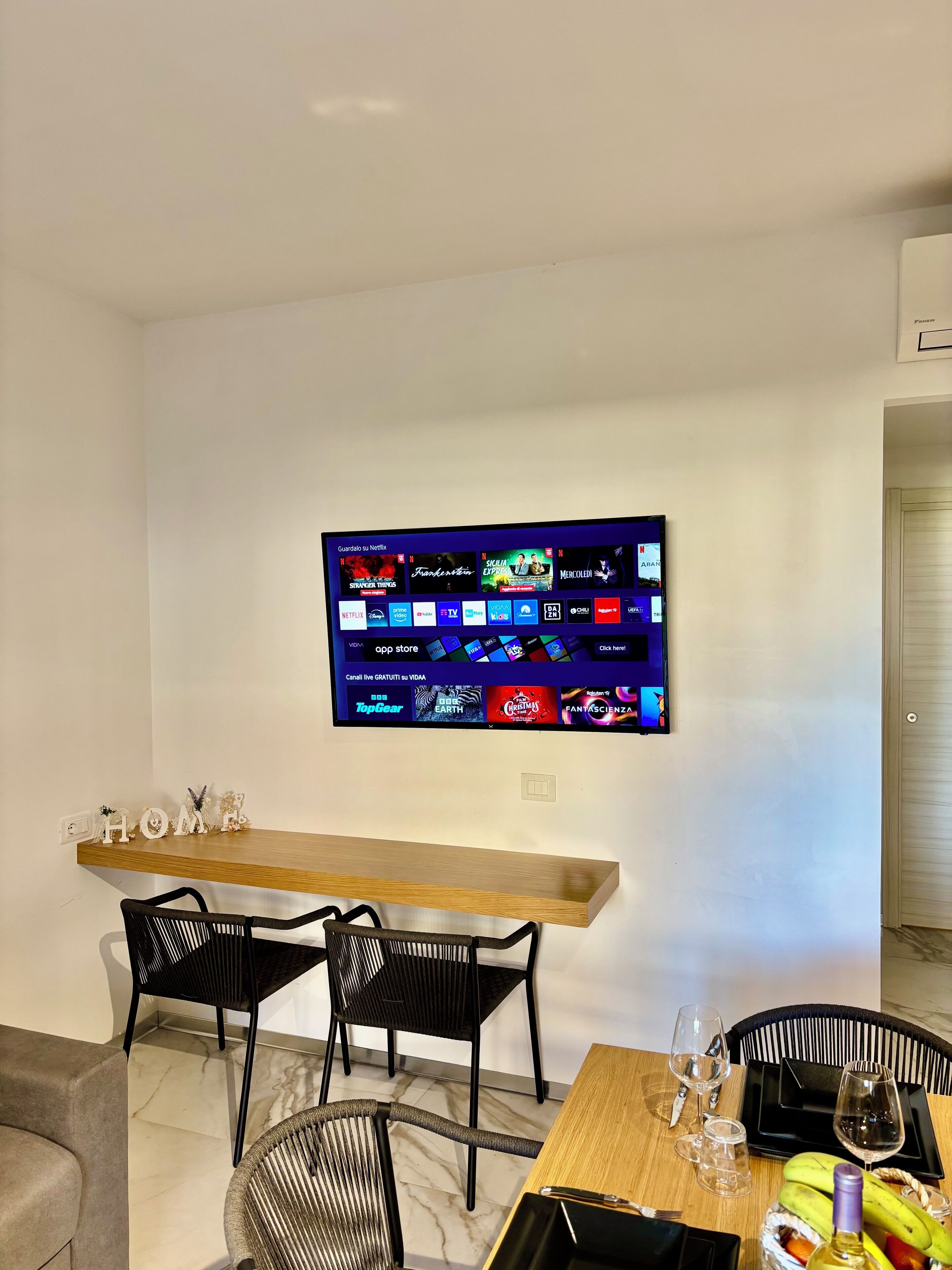 Una sala moderna con un televisor de pantalla plana montado en la pared, mostrando varios servicios de transmisión. Hay dos sillas negras y una mesa de comedor de madera con algunos objetos encima, incluyendo una botella y vasos.