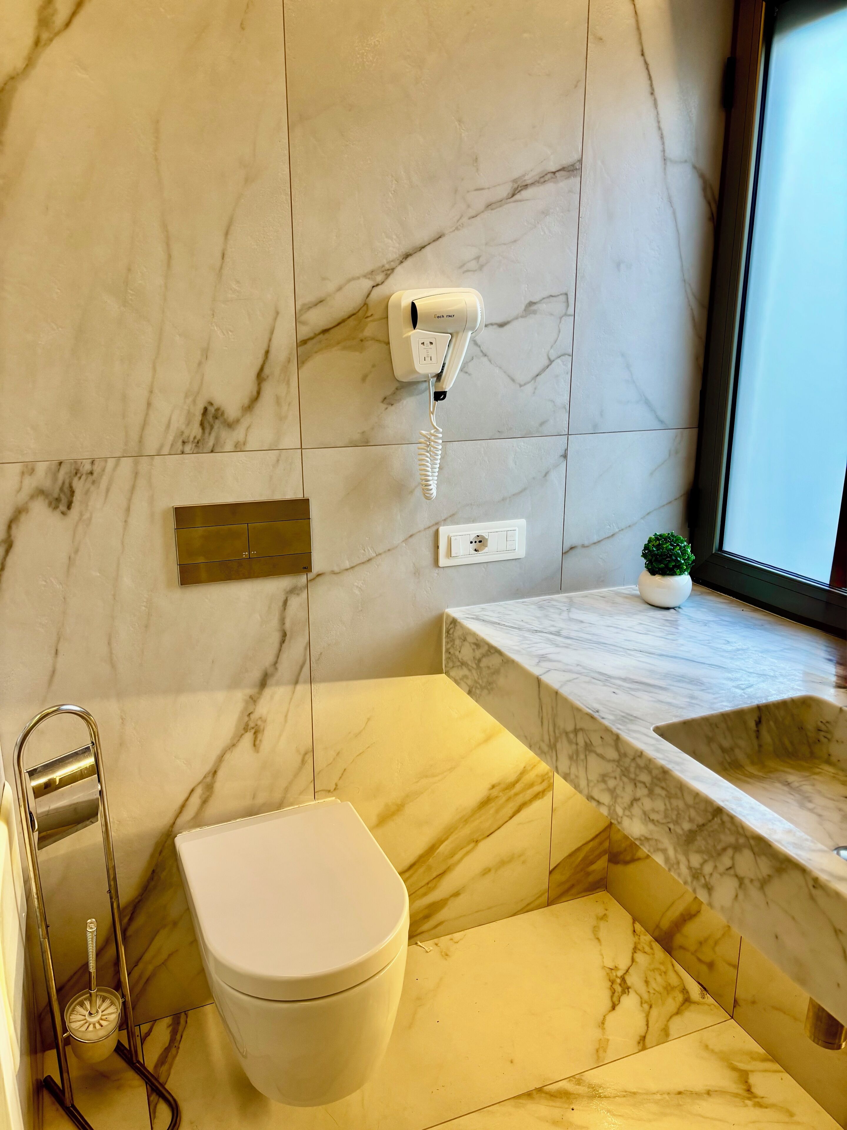 A bathroom with a marble floor, a toilet, a sink with a marble countertop, a hairdryer mounted on the wall, and a small potted plant on the countertop.