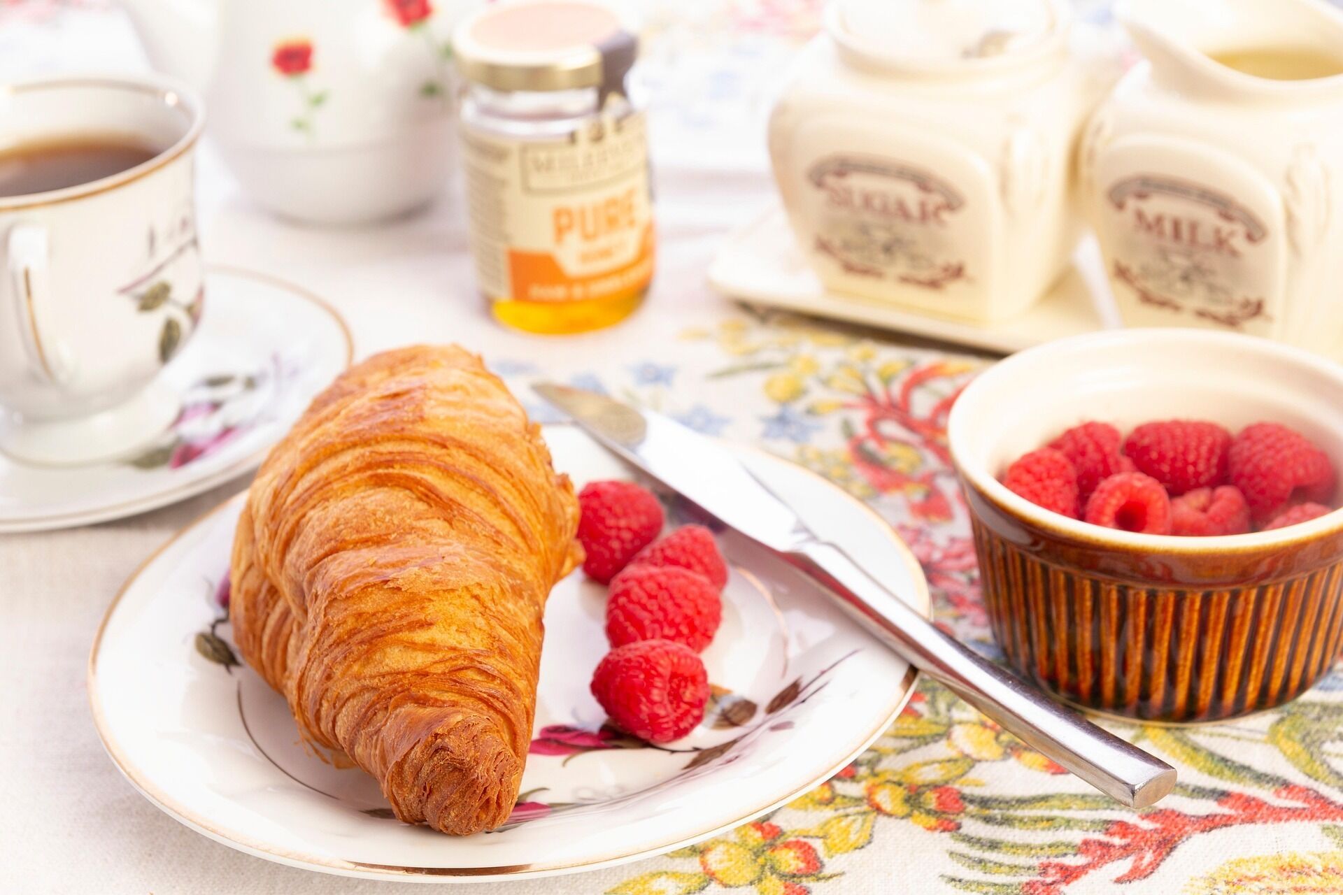 Un croissant sobre un plato con un cuchillo, un bol de frambuesas y botes de leche y mantequilla sobre una mesa.