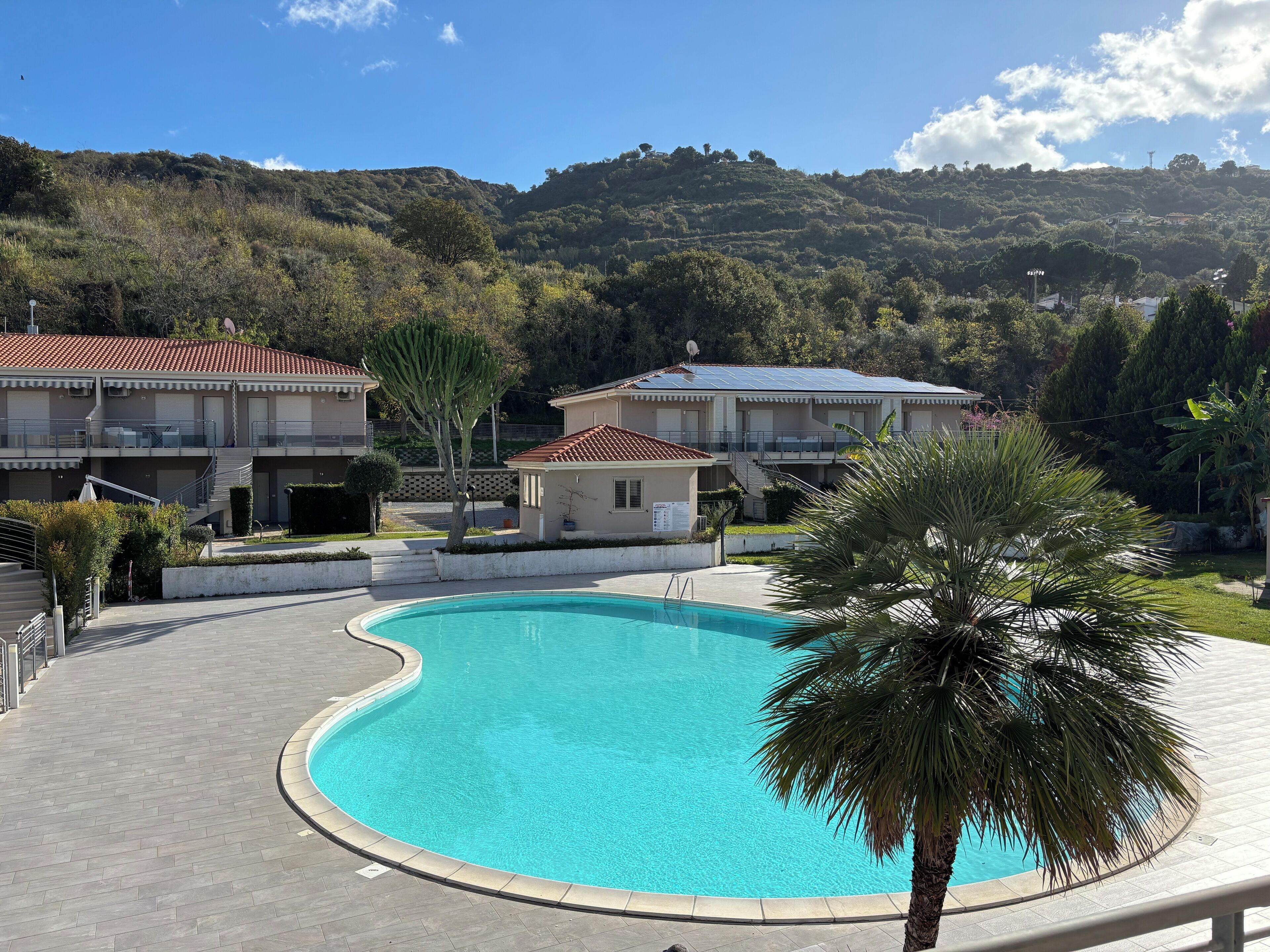 Una piscina con bordo curvo, circondata da un vialetto lastricato. Si vedono edifici con tetti in tegole rosse e balconi, e una collina con alberi sullo sfondo.