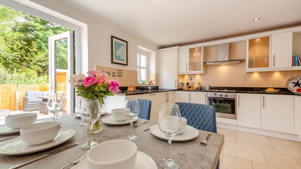 A modern kitchen with white cabinets, a dining table set with white plates and glasses, and a vase of pink flowers. - 4 Bed in Abersoch (oc-b29438) (Abersoch)