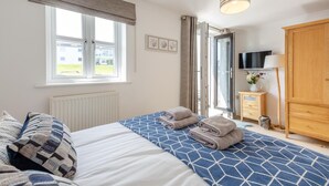 A bedroom with a bed, a window, a TV, a wardrobe, and a bedside table with a lamp and a vase of flowers. - 4 Bed in Abersoch (oc-b29438) (Abersoch)