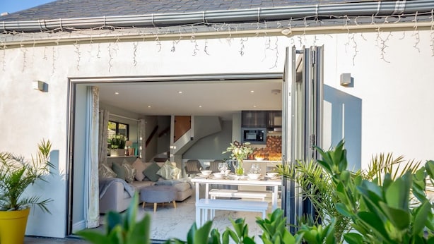 Ein modernes Haus mit einer großen, offenen Terrasse, einem Essbereich und einer durch eine offene Tür einsehbarer Küche.