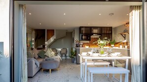 Ein modernes Wohnzimmer mit einer Couch und einem Hocker, ein Essbereich mit Tisch für vier Personen und eine Küche mit dunklen Schränken und einer Spritzschutzwand.