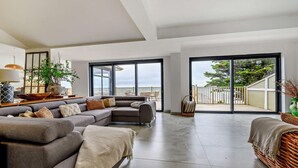 A spacious living room with a grey sectional sofa, a coffee table, and a large window offering a view of the outdoors. - 3 Bed in Downderry (oc-s29364) (Downderry)