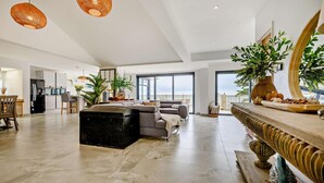A spacious living area with a large sofa, a black ottoman, and a dining table with a decorative centerpiece. There are wicker pendant lights and a large window offering a view of the outdoors. - 3 Bed in Downderry (oc-s29364) (Downderry)