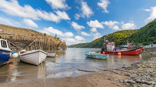Un paysage côtier avec des bateaux amarrés à un quai en pierre, une plage de sable et une colline couverte de végétation.