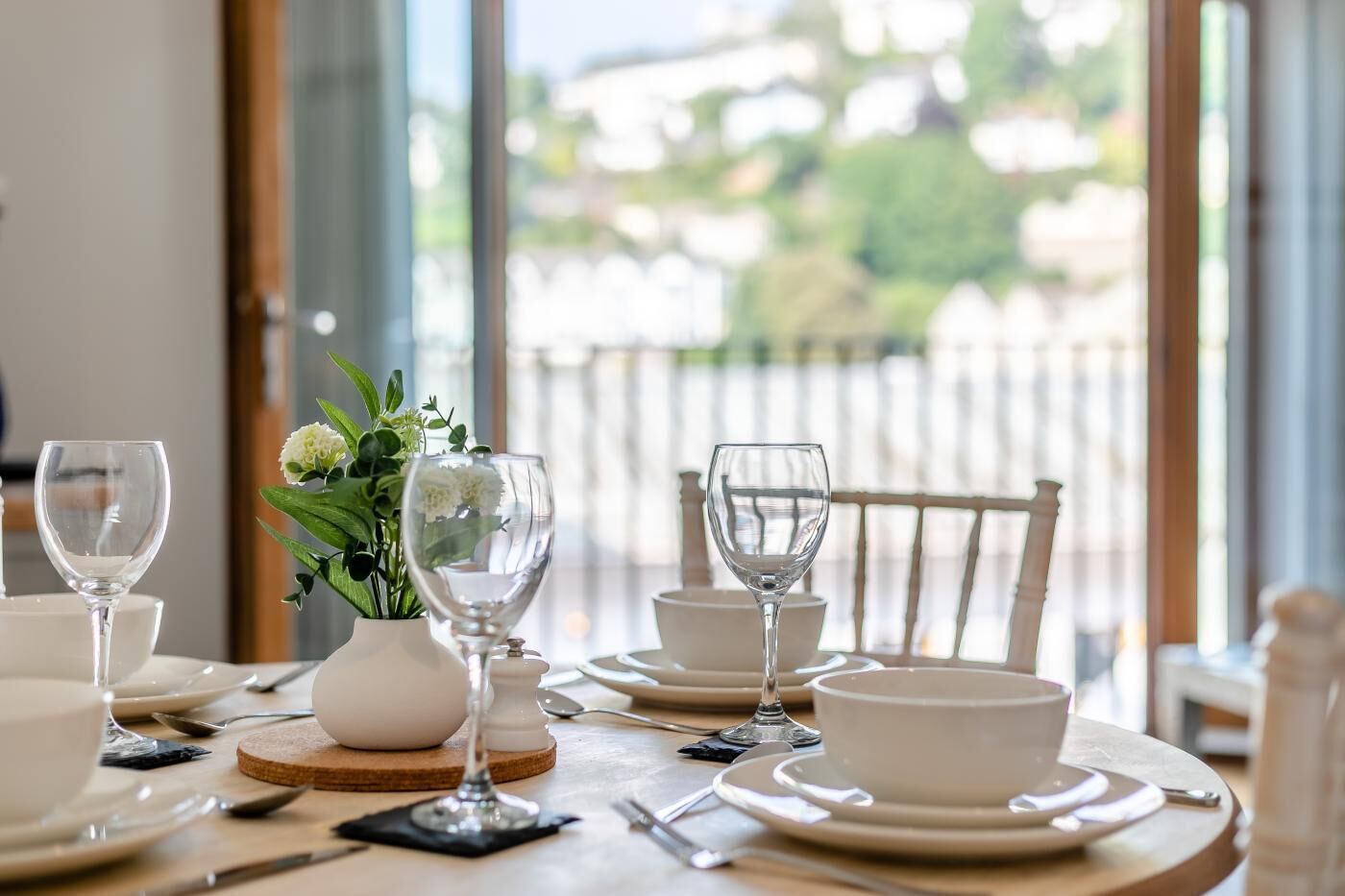 Mesa de jantar posta com pratos, tigelas e copos de vinho brancos, com um vaso de flores e vista para o verde através de janelas amplas.