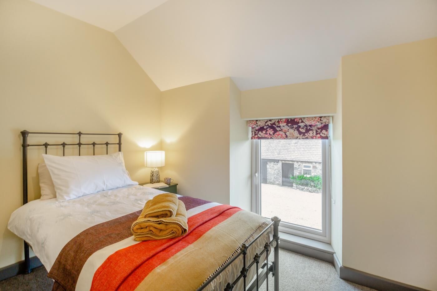 A bedroom with a bed, a nightstand, a lamp, and a window with a view of the outside.
