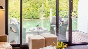 A modern room with a large sliding glass door leading to a balcony overlooking a lake. Inside, there are two cushioned chairs, a small round table with a plant and a tray of pastries, and a wooden floor. - 3 Bed in Looe (oc-j29456) (Looe)