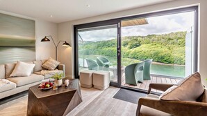 Ein modernes Wohnzimmer mit Sofa, Couchtisch und Sesseln, eine große Schiebeglastür, die auf eine Terrasse mit Seeblick und Waldlandschaft führt.
