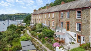 A stone building with a balcony, a garden with a table and chairs, and a view of boats on the water.