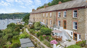 A stone building with a balcony, a garden with a table and chairs, and a view of boats on the water.