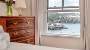 A bedroom with a wooden dresser, a lamp, and a view of a coastal town through a window.