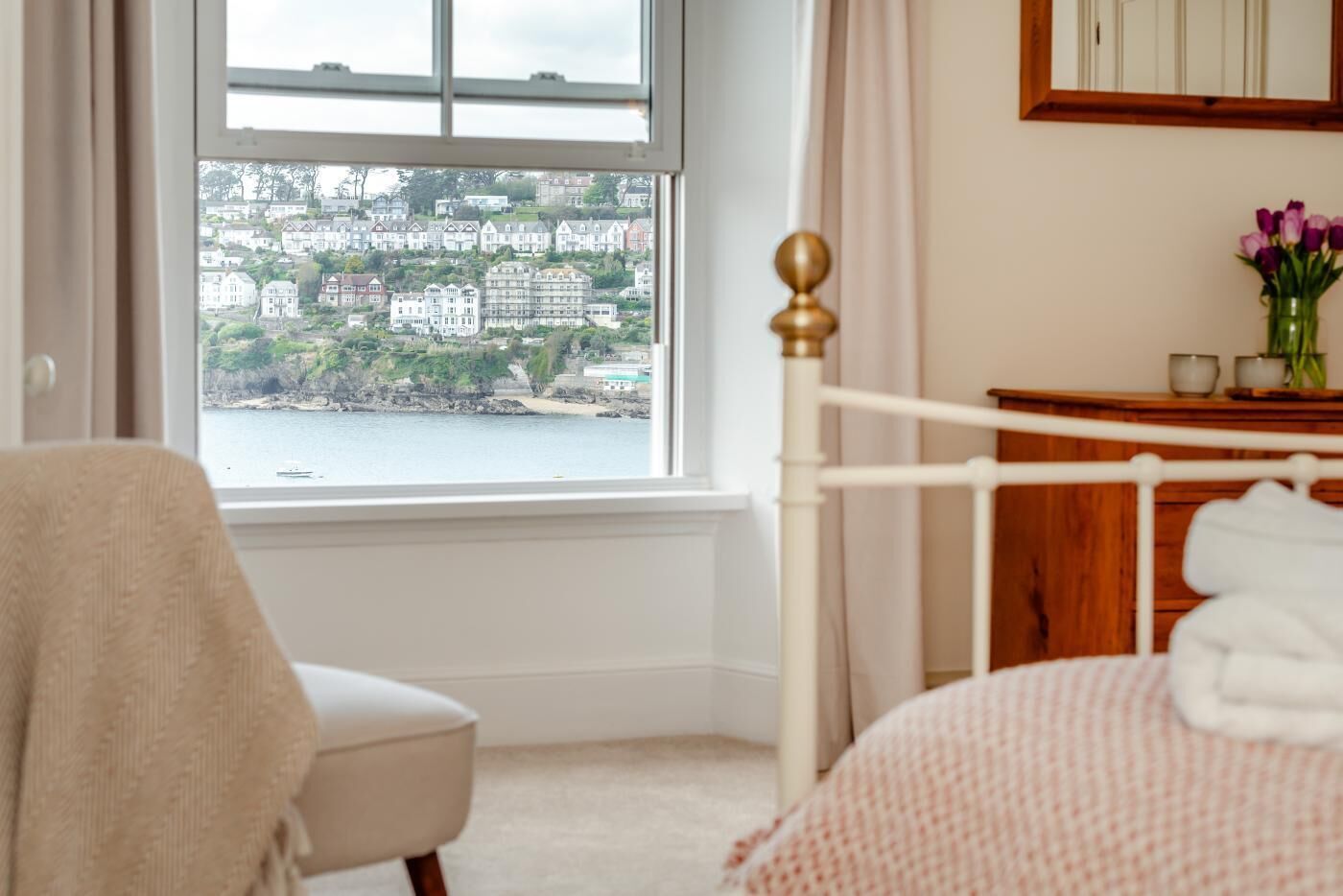 A bedroom with a view of a coastal town, a wooden bed frame, a chair, and a bedside table with a vase of flowers.