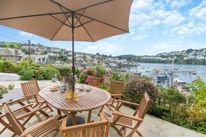 A wooden table with chairs and an umbrella, overlooking a harbor with boats and houses. - 4 Bed in Polruan (oc-n29511) (Fowey)