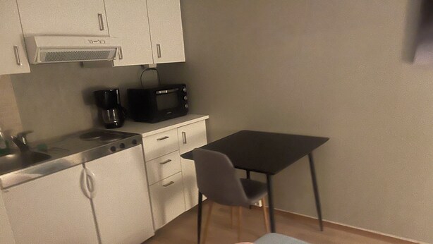 Een kleine keuken met een magnetron, koffiezetapparaat en een tafeltje met een stoel.