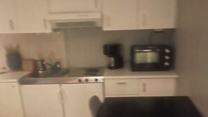 Een kleine kitchenette met een magnetron, koffiezetapparaat en een spoelbak. Er is een zwart tafel en een bed met een roze deken.
