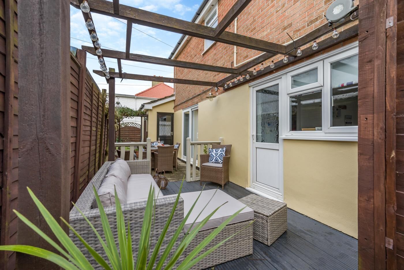 Ein Patio mit Rattan-Sofa, Esstisch und Stühlen. Eine Holz-Pergola mit hängenden Lichtern ist vorhanden.
