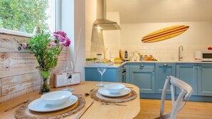 Une cuisine moderne avec des meubles bleus, une table à manger en bois pour deux personnes et une grande fenêtre donnant sur un espace verdoyant.