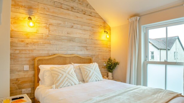 Une chambre à coucher avec une tête de lit en bois, deux lampes de chevet, une table de chevet avec un plateau, et une fenêtre donnant sur des maisons.