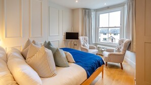 A bedroom with a bed, a TV, a chair, and a view of buildings outside the window. - 6 Bed in Abersoch (oc-c29569) (Abersoch)