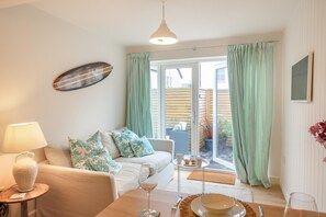 A cozy living room with a sofa, a coffee table, and a dining table set for two. A large window with curtains allows natural light to fill the space. - 1 Bed in Abersoch (oc-w29568) (Abersoch)