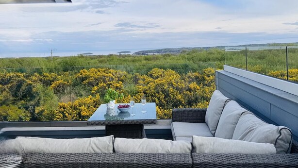 A rooftop terrace with a wicker sofa, a glass table with a bowl of fruit and wine glasses, and a view of a coastal landscape. - 3 Bed in Mynytho (oc-m29517) (Mynytho)