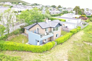 A modern two-story house with a balcony, surrounded by well-maintained hedges and a grassy lawn. - 3 Bed in Mynytho (oc-m29517) (Mynytho)