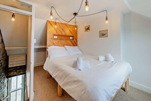 A bedroom with a wooden headboard, a bed with white linens, and a string of exposed bulbs on the ceiling. - 2 Bed in Looe (oc-a29612) (Looe)