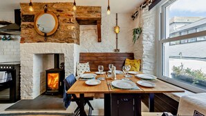 A cozy dining area with a stone fireplace, wooden table, and a view of the city through large windows. - 2 Bed in Looe (oc-a29612) (Looe)