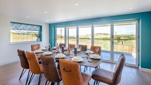 A dining room with a large table set for six, large windows with a view of the countryside, and a balcony with a wooden railing. - 5 Bed in Aberdaron (oc-b29652) (Anelog)