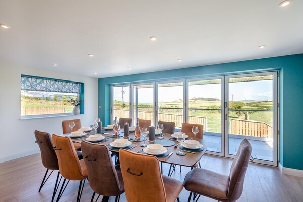 Een eetkamer met een grote tafel gedekt voor zes personen, grote ramen met uitzicht op het landschap, en een balkon met een houten balustrade.