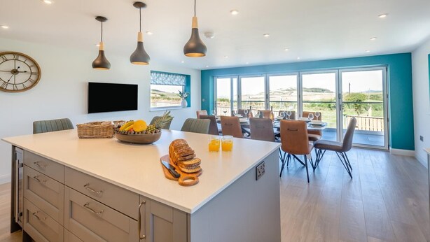 A modern kitchen with a large island, dining area with chairs, and a view of the outdoors. - 5 Bed in Aberdaron (oc-b29652) (Anelog)