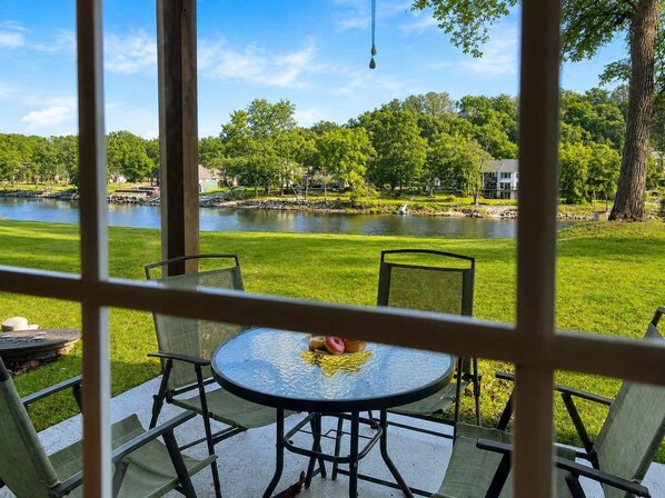 A glass-enclosed patio with a round table and chairs, overlooking a lake and a residential area. - 692 Walk-in Lakefront Oasis - Cozy, Calm, Scenic (Branson)