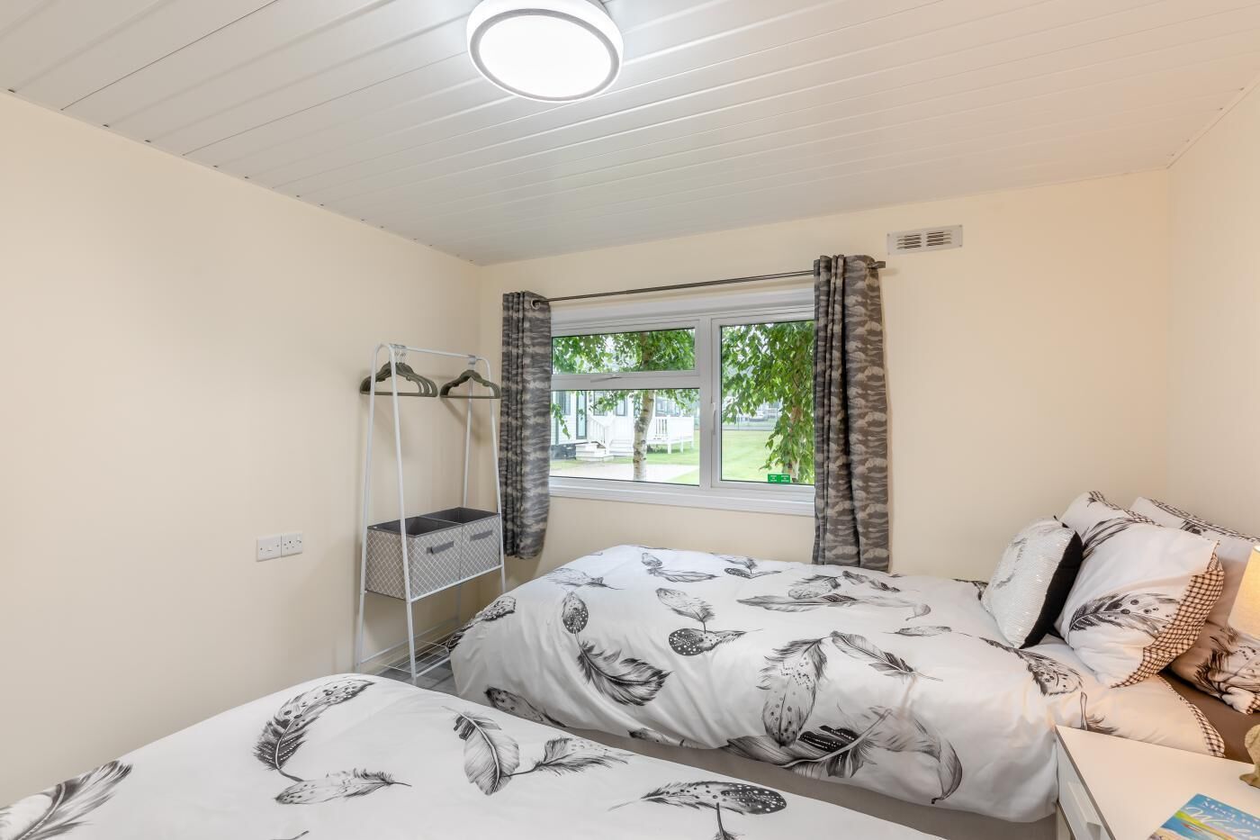 A bedroom with two beds, a window with curtains, a bedside table, and a hanging storage unit.