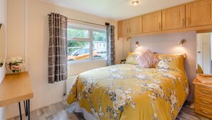 A bedroom with a bed, a window with curtains, a wooden cabinet above the bed, and a small table with a vase of flowers.