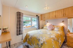 A bedroom with a bed, a window with curtains, a wooden cabinet above the bed, and a small table with a vase of flowers.