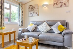 A grey leather sofa with floral cushions, a wooden coffee table with a mug and a book, and a window with curtains.