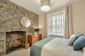 Ein Schlafzimmer mit Kamin aus Stein, einem Bett mit blauen Kissen, einem Nachttisch mit Lampe und einem Fenster mit Jalousien.