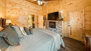 Un dormitorio de cabaña de madera con una cama, almohadas, un ventilador de techo, una chimenea, un televisor y paredes de madera.