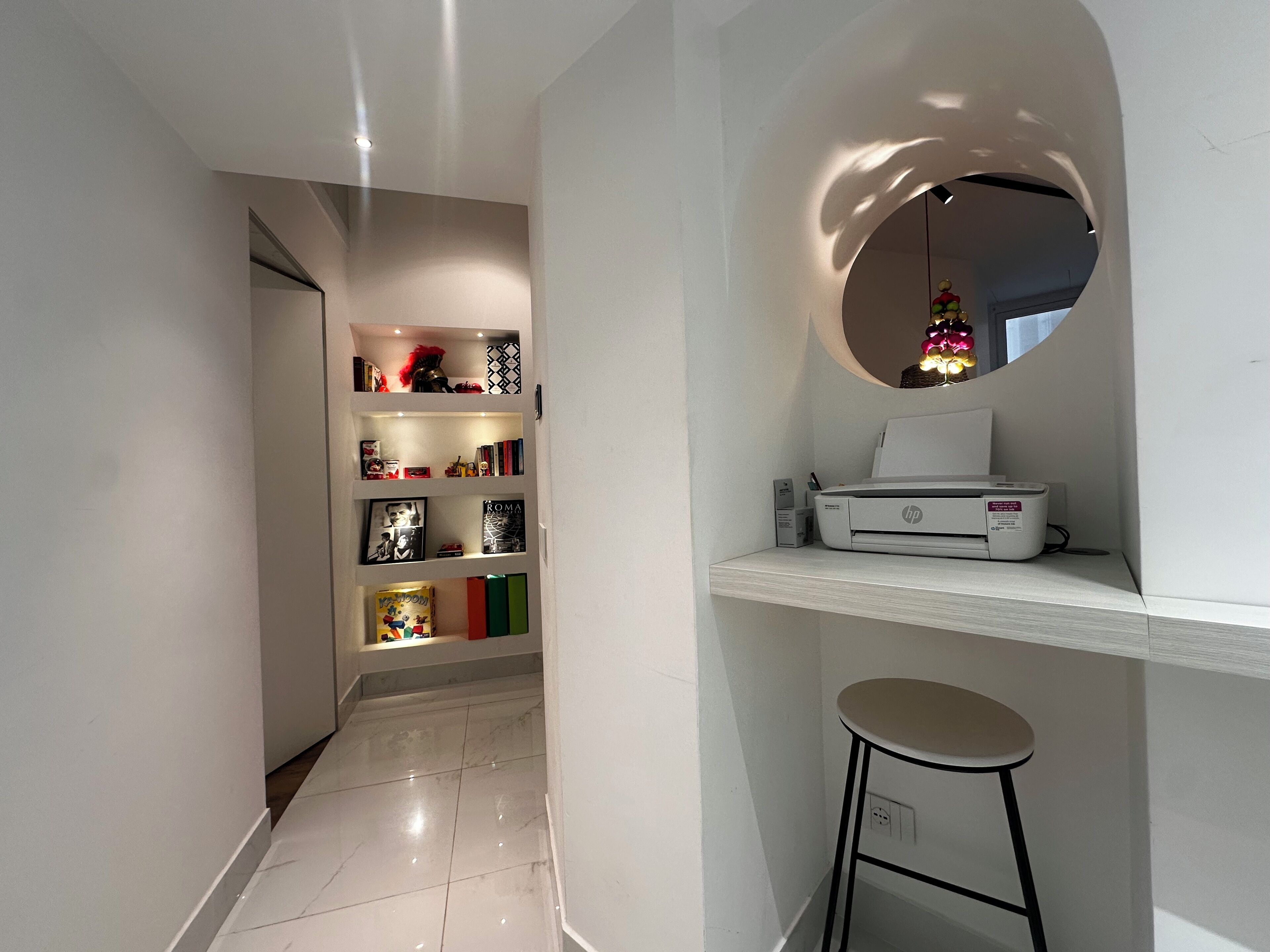 A modern hallway with a white wall, a bookshelf with various items, a printer, and a round mirror with a Christmas ornament.