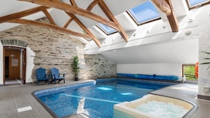 Una piscina interna moderna con parete in pietra e travi in legno, una vasca idromassaggio e due sedie blu.