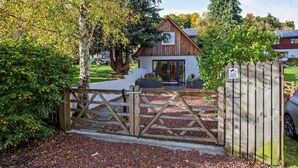 A wooden gate leading to a house with a modern design, a paved driveway, and a sign indicating CCTV surveillance. - 1 Bed in Strathpeffer (oc-h30077) (Strathpeffer)