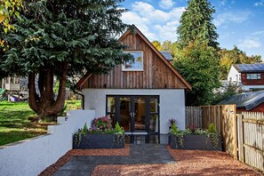 Una pequeña casa de madera con diseño moderno, un gran árbol en la entrada y un patio trasero cercado.