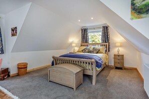 A bedroom with a sloped ceiling, a bed with a wooden headboard, two bedside tables with lamps, a small wooden chest, and a window with curtains. - 1 Bed in Strathpeffer (oc-h30077) (Strathpeffer)