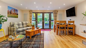 A living room with a sofa, coffee table, and dining table with chairs. There is a television mounted on the wall and a window with curtains. - 1 Bed in Strathpeffer (oc-h30077) (Strathpeffer)