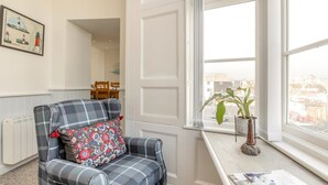 A cozy room with a plaid armchair, a radiator, a window with a view of the city, and a table with a plant and a magazine. - 2 Bed in Penzance (oc-g30154) (Penzance)